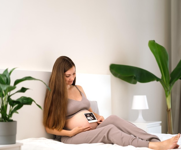 Beautiful caucasian brunette pregnant woman sitting on bed looking at ultrasound photosonogram strok