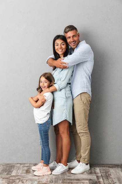 beautiful caucasian family woman and man with little girl smiling and hugging together, isolated over gray