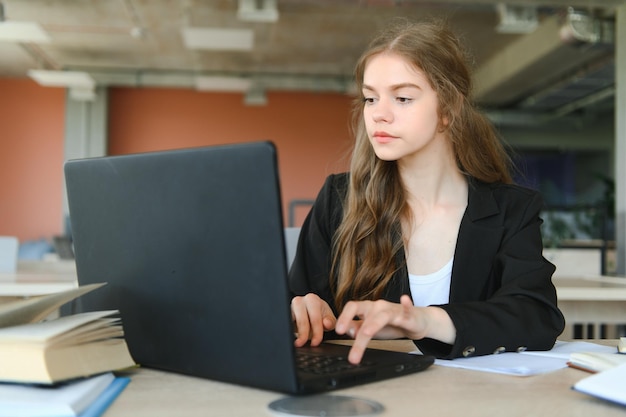 A beautiful caucasian female student is studying in college remotely She is sitting with a laptop and a notepad and concentrated is watching a video conference lesson