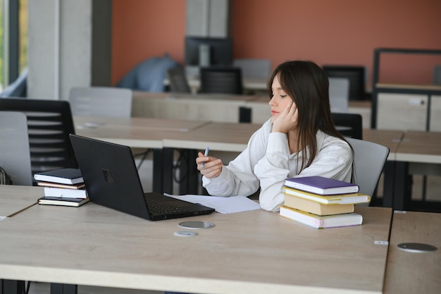 A beautiful caucasian female student is studying in college remotely She is sitting with a laptop and a notepad and concentrated is watching a video conference lesson