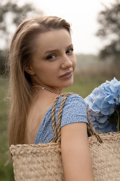 Beautiful caucasian girl in a blue dress walks through the field with a basket of flowers and looks at the camera