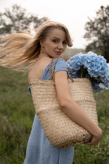 A beautiful caucasian girl in a blue dress and with her hair flying in the wind walks through the field with a basket of flowers and looks at the camera