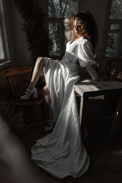 Beautiful caucasian girl bride with curly red hair sits in a wedding dress on the table in a fashion pose