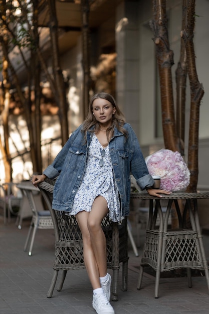 A beautiful caucasian girl in a casual outfit sits in a street cafe with a bouquet of beautiful peonies on the background and looks at the camera