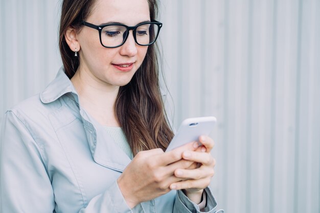 Beautiful caucasian girl in a gray coat and glasses type a message on a smartphone