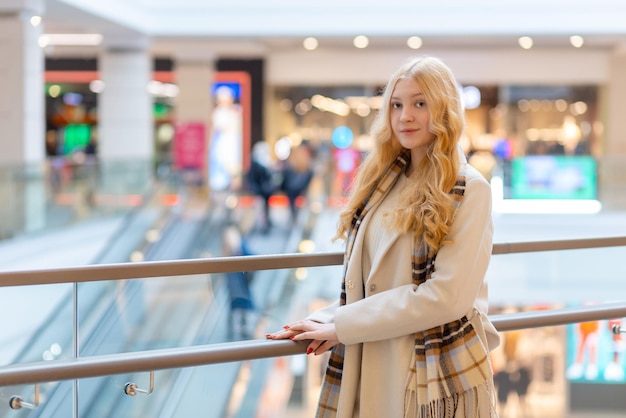 Beautiful caucasian girl in light beige coat poses near glass enclosure mall