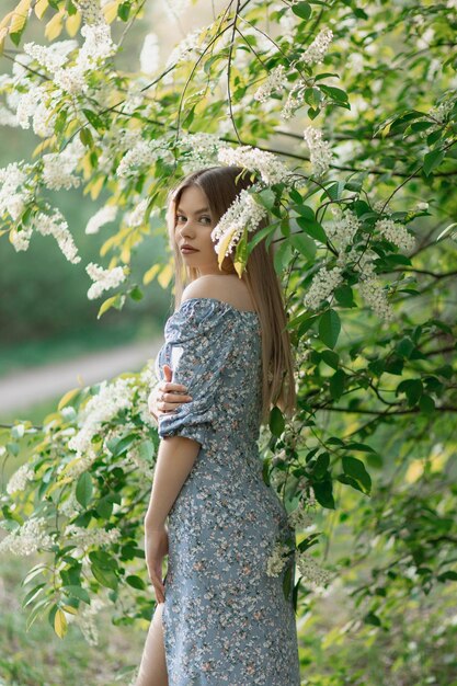 Una bella ragazza caucasica sta in un vestito blu vicino a un albero in fiore e guarda la telecamera