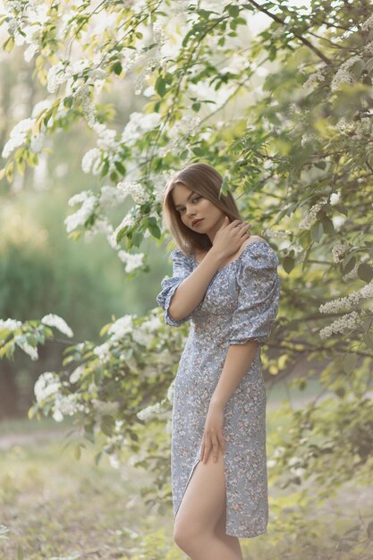 A beautiful caucasian girl stands in a blue dress near a flowering tree and looks at the camera