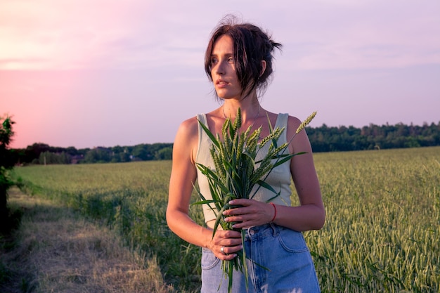 Красивая кавказская девушка в пшеничном поле с букетом пшеничных колосьев. Европейская девушка на поле в лучах заката.