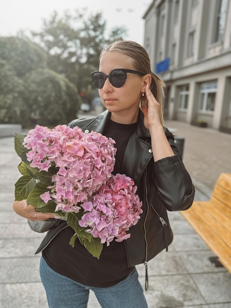 Beautiful caucasian girl with blond hair in a black jacket and blue jeans with a bouquet of pink hydrangeas and with the sunglasses with a city view in the background