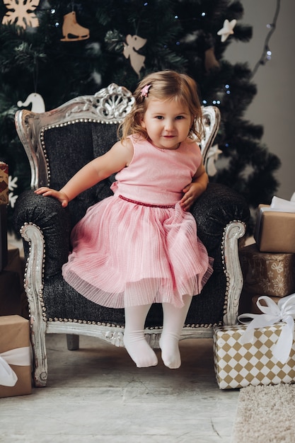 A beautiful caucasian girl with short blonde wavy hair in a cute pink dress rejoices near the Christmas tree and a lot of presents in a large light room of the house