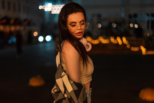 Beautiful caucasian hipster girl with glasses and headphones with a top and shirt walks in the night city with bokeh lights