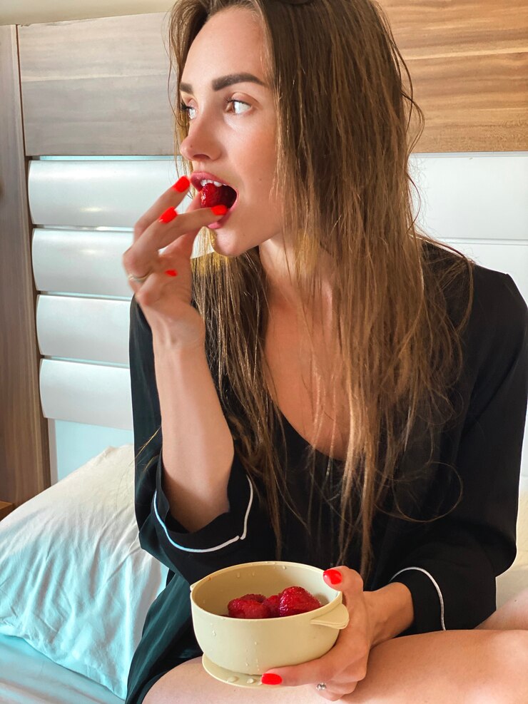 Photo beautiful caucasian lady in hotel bed eats strawberry in black pajamas and looking away