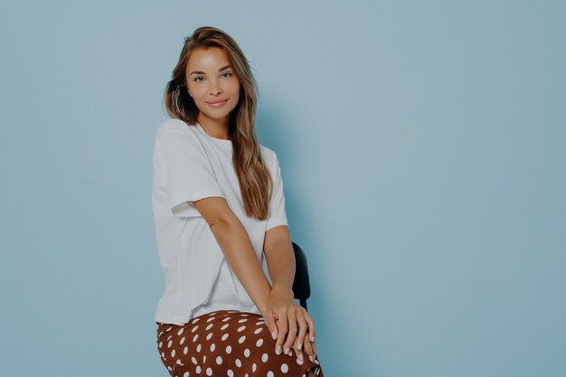 Beautiful caucasian model female sitting on chair with her hands folded on legs with positive face expression, wears casual white tshirt and brown polka dot skirt isolated on blue background