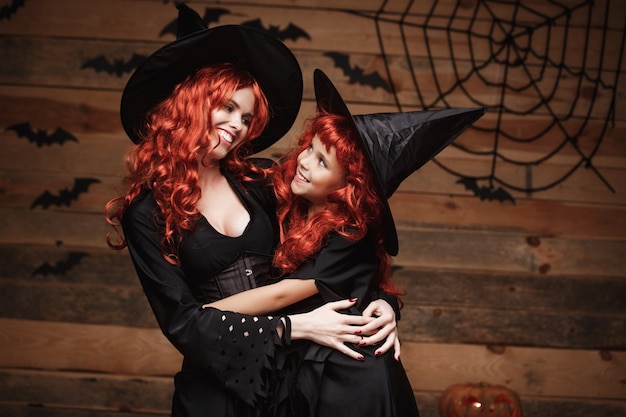 Beautiful caucasian mother and her daughter with long red hair in witch costumes celebrating Halloween posing 