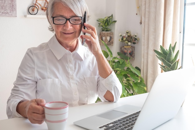 Beautiful Caucasian senior woman in white shirt using laptop while working indoors holding mobile phone in hand smiling lady talking at cellphone