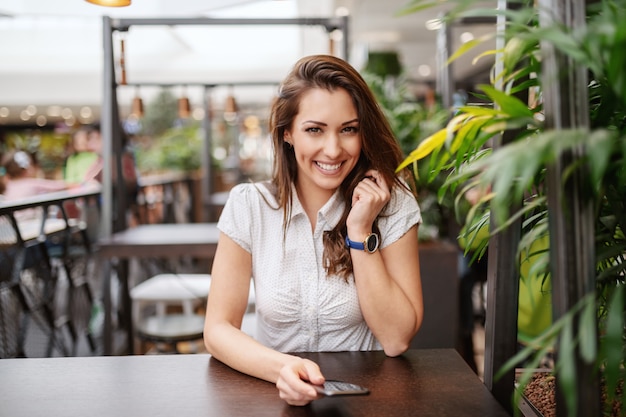 Beautiful Caucasian smiling brunette posing at coffee shop while holding smart phone.