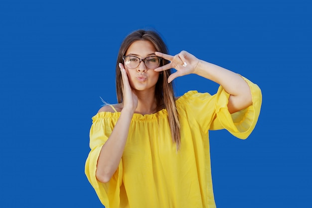 Beautiful Caucasian teenage girl in yellow blouse and with eyeglasses posing in front of blue wall.