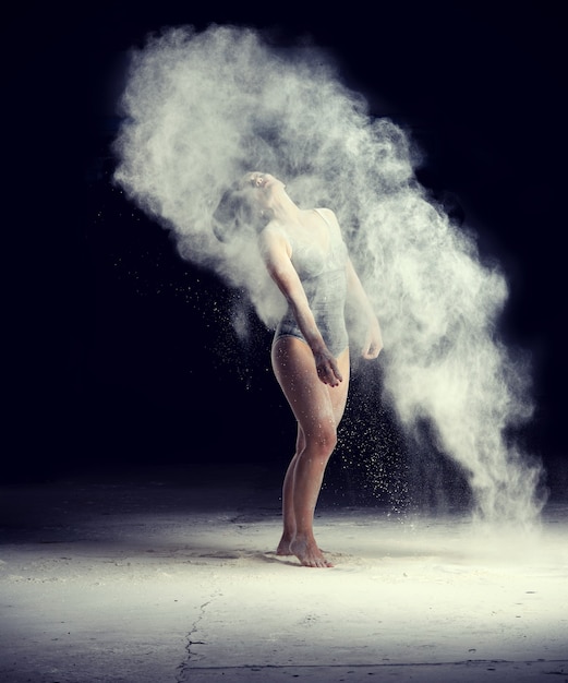 Beautiful caucasian woman in a black bodysuit with a sports figure is dancing in a white cloud of flour on a black background, a white cloud of dust flies out of the hair