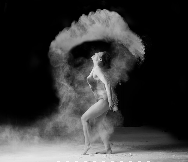 Photo beautiful caucasian woman in a black bodysuit with a sports figure is dancing in a white cloud of flour, black and white toning