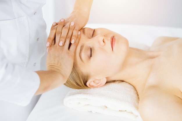 Beautiful caucasian woman enjoying facial massage with closed eyes in sunny spa salon Relaxing treatment in medicine