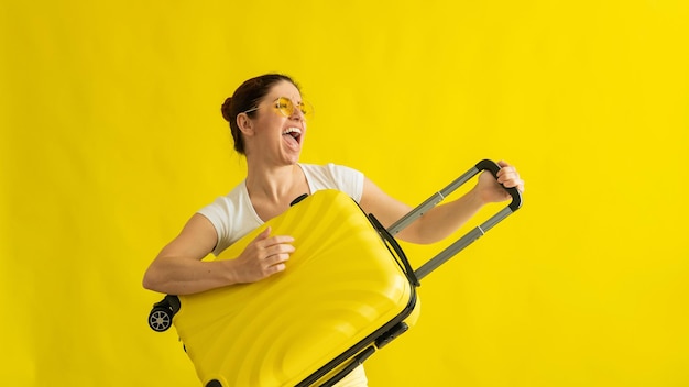 Beautiful caucasian woman fooling around with a suitcase on a yellow background a charming girl imitates playing the guitar the joy of the journey ahead