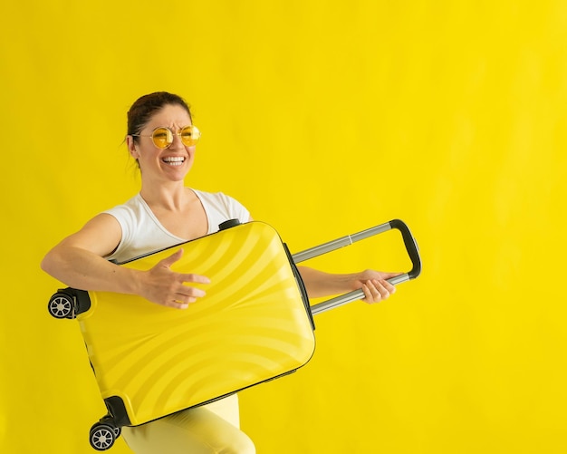Красивая белая женщина шутит с чемоданом на желтом фоне.