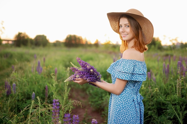 A beautiful Caucasian woman in a hat and dress holds a bouquet and smiles