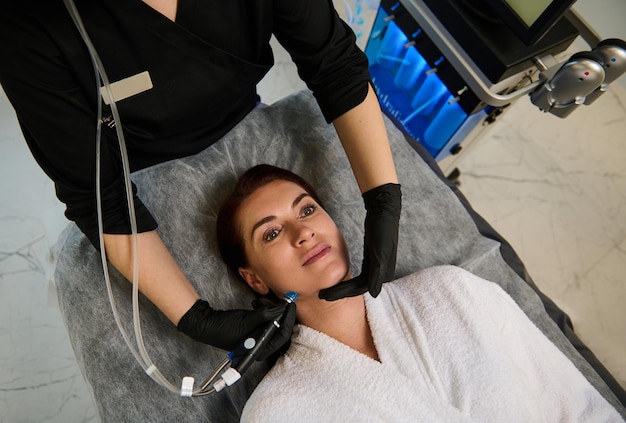 Bella donna caucasica sdraiata su un lettino da massaggio e ricevendo un trattamento viso idro microdermoabrasione per il ringiovanimento della pelle ringiovanimento della terapia antietà cosmetologia hardware