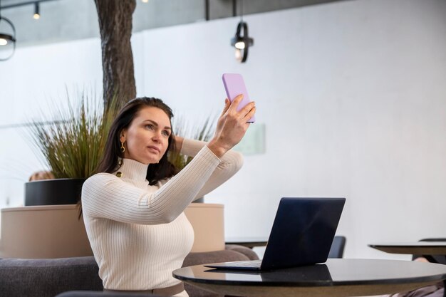 Bella donna caucasica fa selfie su uno smartphone seduto a un tavolo con un laptop un blogger