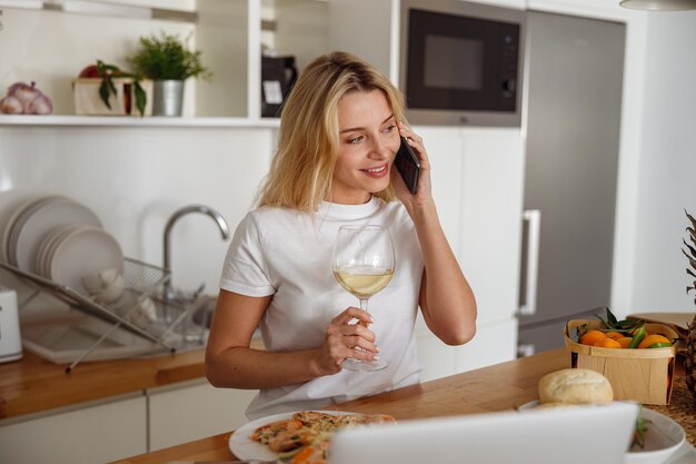 Beautiful Caucasian woman talking on cellphone and having white wine in glass in kitchen