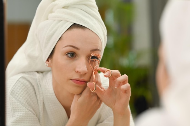 Photo beautiful caucasian woman using eyelash curler curling eyelashes before mascara in front of mirror