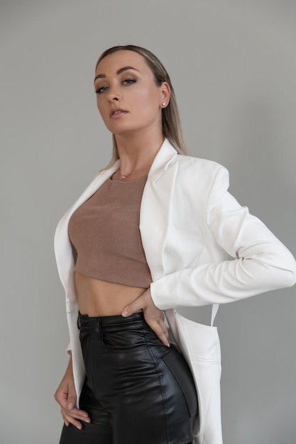 A beautiful caucasian woman in a white jacket and black trousers stands in a fashion pose in the studio on a gray background and looks at the camera