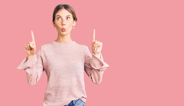 Beautiful caucasian woman with blonde hair wearing casual winter sweater amazed and surprised looking up and pointing with fingers and raised arms