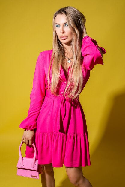 Beautiful caucasian woman with long fair hair in pink dress holding a small bag and posing to the camera on bright yellow background