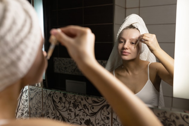 Bella donna caucasica con un asciugamano in testa in bagno dopo una doccia si guarda allo specchio