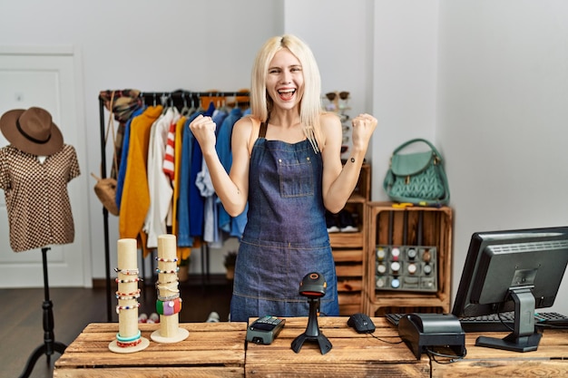 Beautiful caucasian woman working as manager at retail boutique screaming proud celebrating victory and success very excited with raised arms