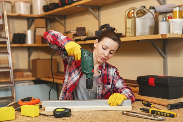 Beautiful caucasian young brown-hair woman in plaid shirt and gray T-shirt working in carpentry workshop at table place, drilling with drill holes in piece of iron and wood while making furniture.