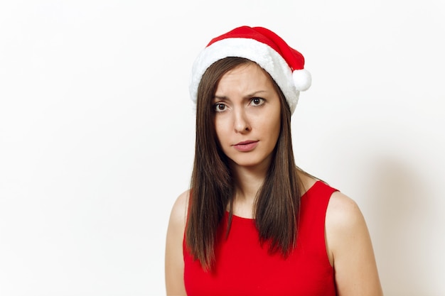 The beautiful caucasian young displeased and discouraged woman wearing red dress and Christmas hat on white background. Santa girl isolated portrait. New Year holiday 2018 concept