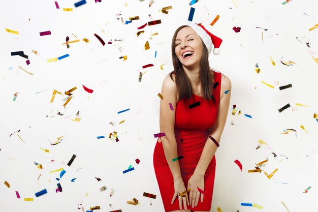 The beautiful caucasian young happy woman wearing red dress and Christmas hat laughing with her eyes closed with pleasure on white background with confetti. Santa girl. New Year holiday 2018 concept