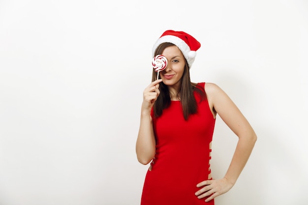 하얀 배경에 사탕을 들고 있는 크리스마스 모자와 빨간 드레스를 입고 건강한 피부와 매력적인 미소를 가진 아름다운 백인 젊은 행복한 여성. 고립 된 산타 소녀입니다. 새해 휴일 2018 개념