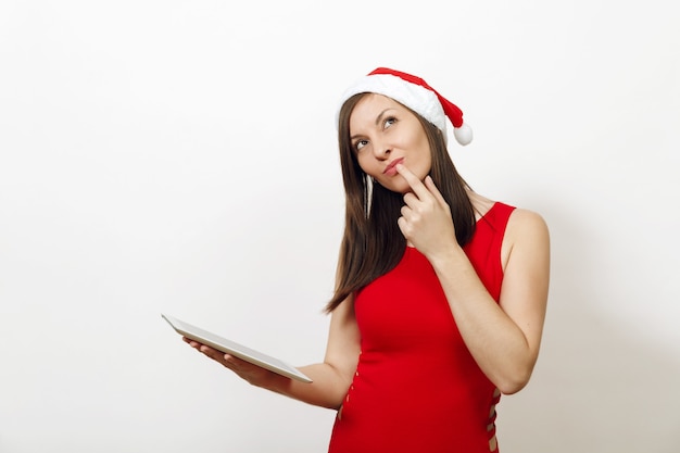 Beautiful charming caucasian young happy woman in red dress and Christmas hat holding tablet on white background. Santa girl and gadget isolated. New Year holiday 2018. Copy space for advertisement.