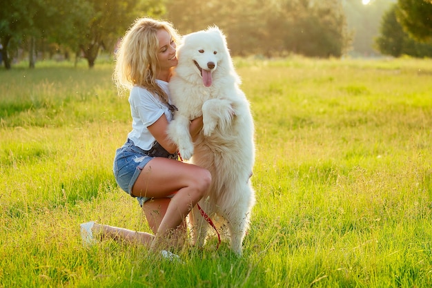 Beautiful and charming curly blonde smiling toothy woman in denim shorts are sitting at glass hugging white fluffy cute samoyed dog in the summer park sunset rays field background . pet and mistress.