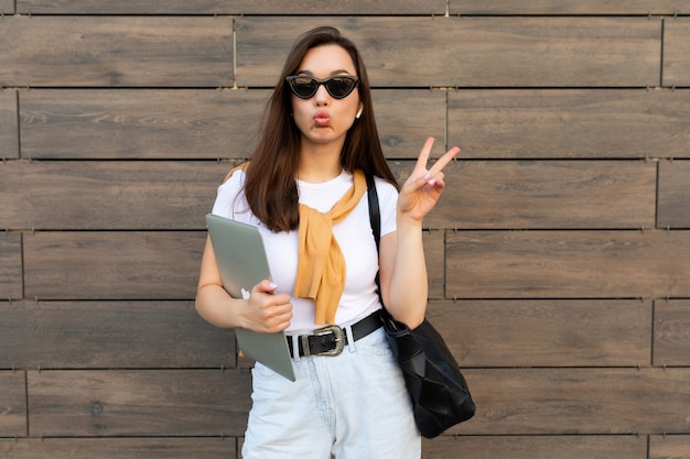 Beautiful charming young brunet woman looking at camera holding computer laptop and sunglasses showing peace gesture with kissing lips in white t-shirt and light blue jeans in the street.