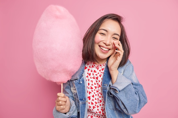 아름 다운 쾌활 한 아시아 여자는 친구와 함께 주말을 보낸다 얼굴에 손을 유지 유행 옷을 입고 행복을 표현 핑크 벽에 고립 된 막대기에 사탕 치실을 보유