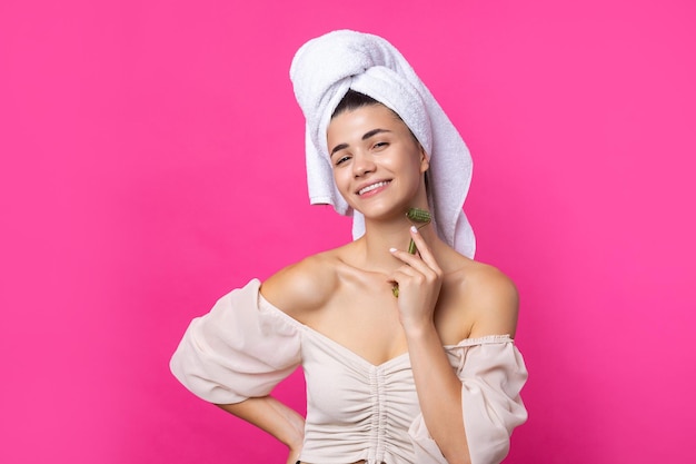 A beautiful cheerful attractive girl with a towel on her head holds a cosmetic roller near her face against a pink background