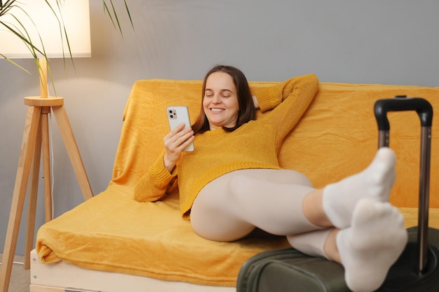 오렌지색 스웨터를 입은 아름다운 쾌활한 백인 여성이 소파에 짐을 들고 앉아 웃으며 휴대폰 화면을 보고 호텔을 떠나기 전에 인터넷을 검색하고 있습니다.