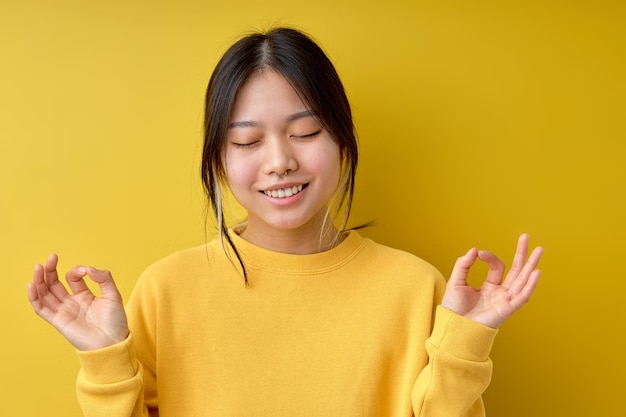 사진 노란색 외진 배경 위에 명상을 하는 아름다운 중국 여성은 긴장을 풀고 손가락으로 명상 제스처를 하며 눈을 감고 미소를 지으며 요가 개념 초상화 복사 공간