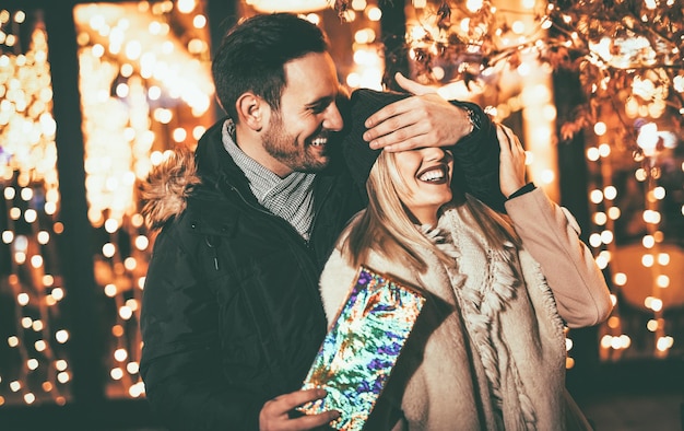 Beautiful cheerful couple celebrating Christmas in the city street. Young man covering the eyes of happy surprised girlfriend and giving her present.