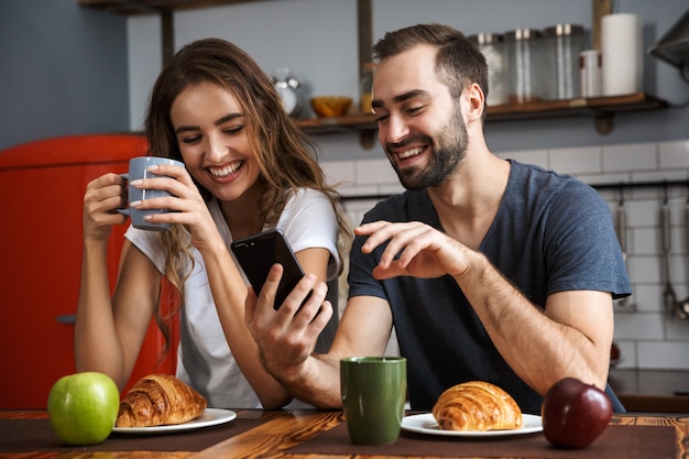 Beautiful cheerful couple having breakfast at the kitchen, using mobile phone